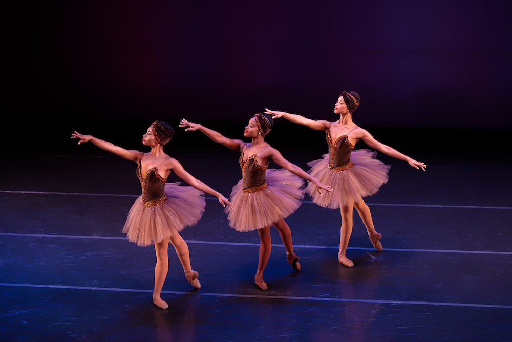 Three Black ballerinas in classical tutus of with crushed velvet and rhinestone bodices and matching crushed velvet head wraps stand in unison, on one leg with the other extended backward on the floor. All different shades, their tights and toe shoes match their various skin tones and they look poised and elegant with their arms out stretched.